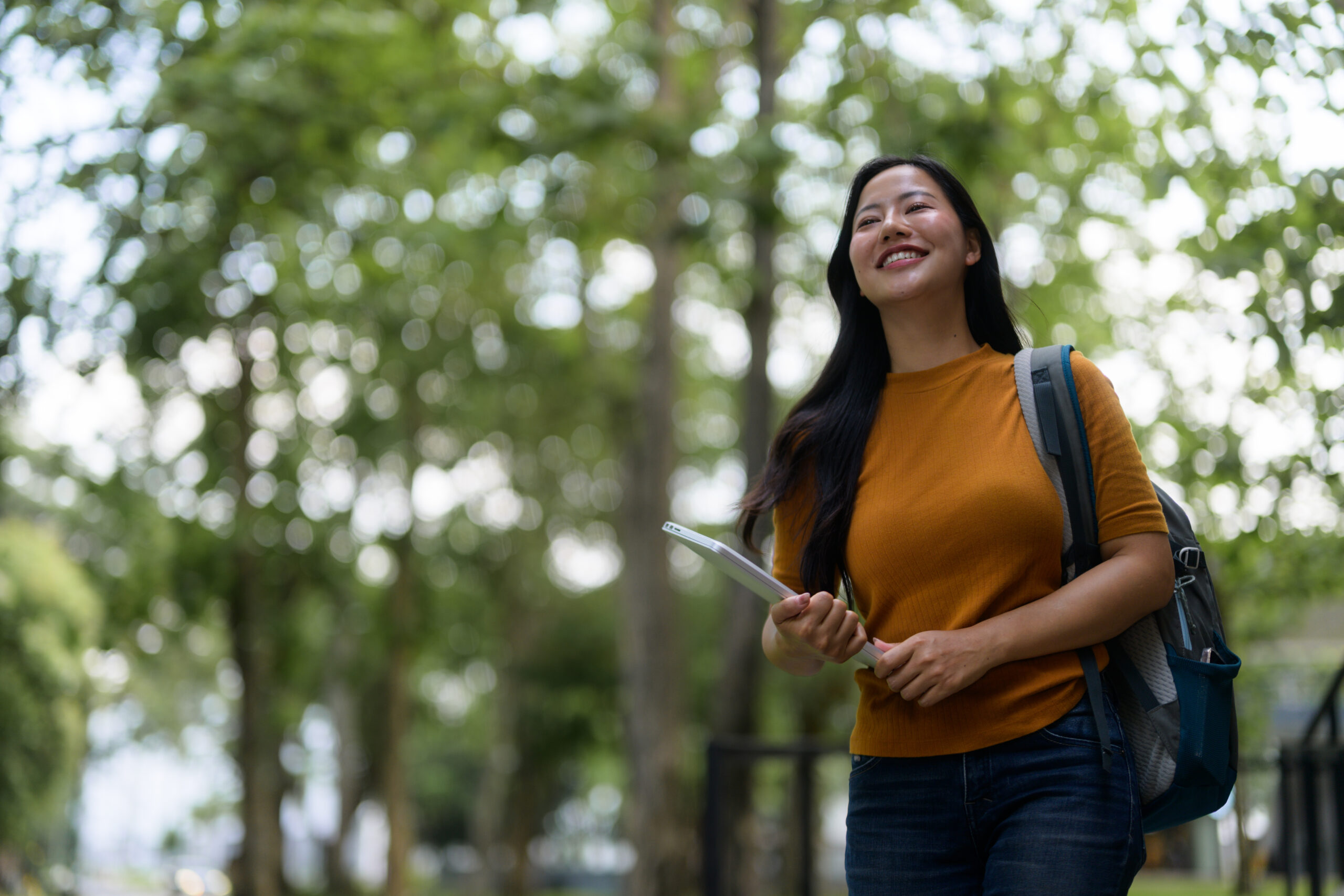 Happy asian student walking in university park holding tablet and backpack, enjoying campus life and looking up